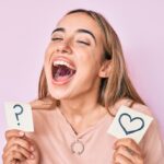 Young beautiful blonde woman holding heart and question mark reminder smiling and laughing hard out loud because funny crazy joke.
