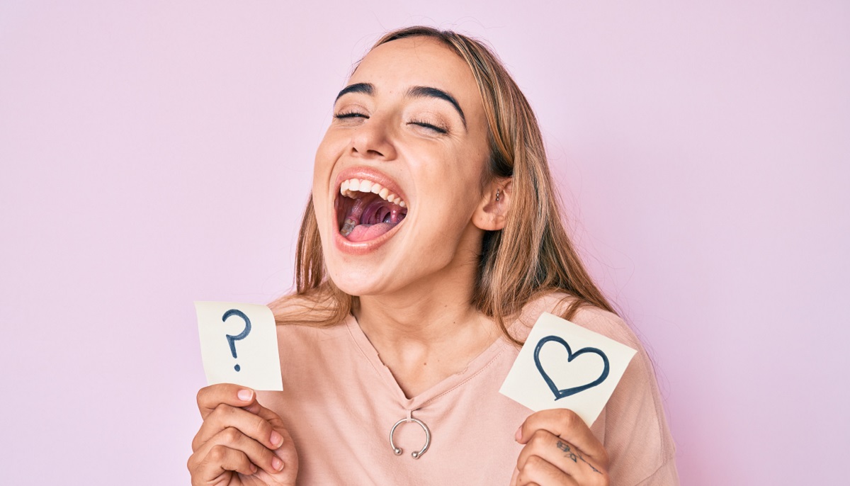 Young beautiful blonde woman holding heart and question mark reminder smiling and laughing hard out loud because funny crazy joke.